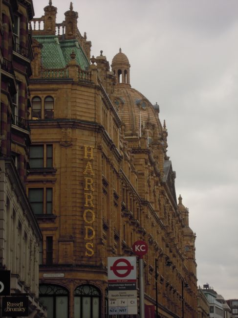 A tall, historic building with ornate architectural details and a domed roof, constructed from honey-colored stone with a textured finish. Several narrow windows with dark frames are visible along the facade, with decorative cornices and spires adorning the upper sections. The building features a vertical illuminated sign reading 'HARRORDS' in yellow letters, positioned on the left side of the structure. In the foreground, there is a red and white London Underground sign indicating nearby public transport, with bus timetable information attached. The scene is set against a cloudy sky, creating a muted, overcast atmosphere, and the building's intricate details and textures contrast with the smooth, modern signage at street level. This urban scene captures a blend of historic architecture and contemporary transportation elements, typical of central London streets where private waste collection services like Waste Disposal Bromley might operate for commercial premises.