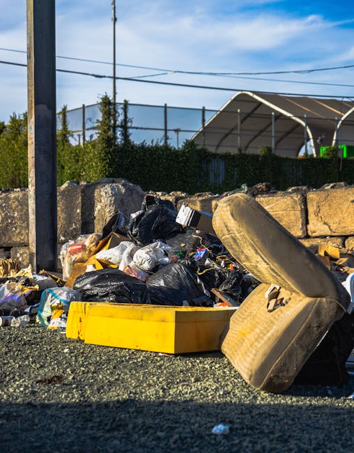 A close-up view of a roadside rubbish pile consisting of black plastic bags filled with waste, various discarded packaging, and an old, worn tire lying on its side. The ground is covered with dark gravel and small debris, with a yellow plastic container or lid partially visible among the waste. Behind the pile are weathered stone blocks forming a low wall, and beyond that, a hedge and some trees. In the background, there are utility poles with overhead power lines and a large, curved industrial-style canopy structure with a metal framework. The scene is illuminated by natural daylight, with a bright blue sky and a few wispy clouds overhead. The image reflects a typical scene of illegal dumping or unmanaged roadside waste, highlighting the importance of professional rubbish removal services offered by Waste Disposal Bromley for private residences or commercial properties seeking alternative waste handling solutions in the Bromley area.