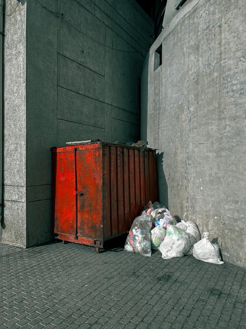 A large, industrial-style red metal skip with visible rust and wear, positioned against a dark green building wall on the left side of the image, and a light grey concrete wall on the right. The skip is situated on a paved surface made of small, dark grey interlocking bricks. To the right of the skip, there are several transparent and white plastic trash bags filled with mixed waste, including identifiable plastic bottles and packaging. The background features a narrow alleyway or passage with shadows cast by the surrounding structures, indicating natural daylight. The setting appears to be an urban or commercial area, typically associated with private waste disposal or on-site clearance services, as provided by Waste Disposal Bromley for rubbish removal needs in Bromley.