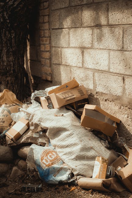 A pile of mixed waste materials, including cardboard boxes, plastic bags, and paper, is stacked against a rough brick and concrete wall outdoors. The cardboard boxes vary in size, some flattened and some partially intact, with visible tape and printed labels. Several plastic bags are crumpled and contain unknown contents, resting on a layer of small rocks and dirt. The scene is shaded by nearby trees or structures, with natural light casting soft shadows, creating a utilitarian environment typical of site clearance or private rubbish disposal areas. The waste collection appears unorganized, indicating a likely case of on-site rubbish accumulation that could be targeted for removal by a local waste management service such as Waste Disposal Bromley, supporting alternative waste handling methods outside of municipal collection systems.