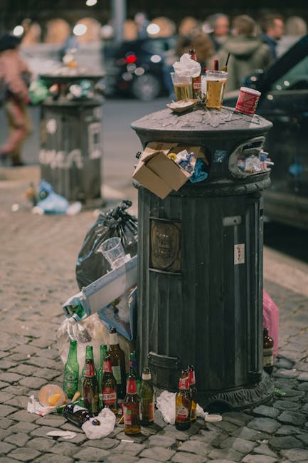 A black, cylindrical metal rubbish bin with a domed lid, situated on a cobblestone pavement in an outdoor urban environment during daytime. The bin is overflowing with discarded items, including fast-food packaging, paper cups, and plastic wrappers, with additional waste surrounding its base such as empty glass bottles, crumpled paper, and plastic bags. Some litter is spilling onto the ground, contributing to the surrounding clutter. In the background, there are blurred figures of pedestrians, parked cars, and other waste bins, indicating a busy street scene. The scene reflects a situation of inadequate waste containment or collection potential, which Waste Disposal Bromley may manage through private rubbish removal or on-site clearance services, ensuring cleaner streets. The overall atmosphere shows a typical scenario of urban litter accumulation, emphasizing the importance of effective waste management to maintain environmental standards in public spaces.